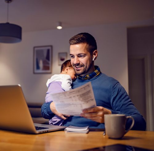 Fatherhood and remote business. A happy young dad works from home holds his sleepy baby girl in his hands, goes through paperwork and has an online meeting on his laptop with his colleagues.